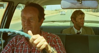 Movie still from “Wake in Fright” (1971), directed by Ted Kotcheff – A man driving a car while another man sits in the passenger seat; Medium shot, Low angle