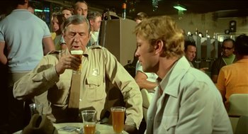 Movie still from “Wake in Fright” (1971), directed by Ted Kotcheff – Two men are drinking beer at a party; Medium shot, Over the shoulder angle