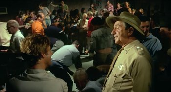 Movie still from “Wake in Fright” (1971), directed by Ted Kotcheff – An older man in a hat is standing next to a crowd of people; Medium shot, Over the shoulder angle