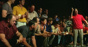Movie still from “Wake in Fright” (1971), directed by Ted Kotcheff – A group of men sitting in a row on a stage; Wide shot, High angle