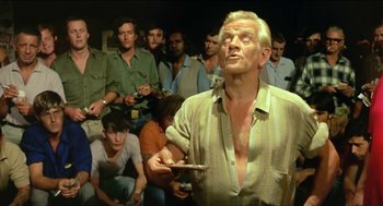 Movie still from “Wake in Fright” (1971), directed by Ted Kotcheff – An old man holding a cell phone in front of a group of people; Medium shot, Over the shoulder angle