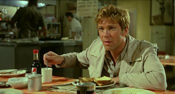 Movie still from “Wake in Fright” (1971), directed by Ted Kotcheff – A man sitting at a table eating a sandwich; Medium shot, Over the shoulder angle