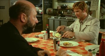 Movie still from “Wake in Fright” (1971), directed by Ted Kotcheff – A group of people sitting at a table eating food; Medium shot, Over the shoulder angle