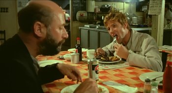 Movie still from “Wake in Fright” (1971), directed by Ted Kotcheff – A group of people sitting at a table eating food; Medium shot, Over the shoulder angle