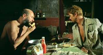 Movie still from “Wake in Fright” (1971), directed by Ted Kotcheff – Two men sitting at a dinner table eating food; Medium shot, Low angle