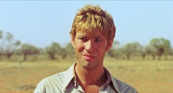 Movie still from “Wake in Fright” (1971), directed by Ted Kotcheff – A man with blonde hair is standing in a field; Close Up shot, Over the shoulder angle
