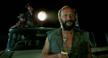 Movie still from “Wake in Fright” (1971), directed by Ted Kotcheff – A man holding a bottle of beer in front of another man; Medium shot, Low angle