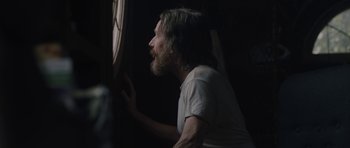 Movie still from “Wakefield” (2016), directed by Robin Swicord – A man with long hair and a beard looking out of a window; Medium shot, Over the shoulder angle