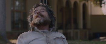 Movie still from “Wakefield” (2016), directed by Robin Swicord – A man with long hair and a beard laying on the ground with his eyes closed; Close Up shot, Low angle