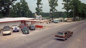 Movie still from “Walking Tall” (1973), directed by Phil Karlson – An old photo of a gas station with cars parked on the side of the road; Extreme Wide shot, High angle