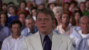 Movie still from “Walking Tall” (1973), directed by Phil Karlson – A man in a white suit and a blue tie in front of a group of people; Close Up shot, Over the shoulder angle