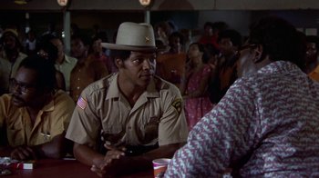 Movie still from “Walking Tall” (1973), directed by Phil Karlson – A man in a hat sitting in front of a group of people; Medium shot, Over the shoulder angle