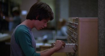 Movie still from “WarGames” (1983), directed by John Badham – A young man is looking at a book on a shelf; Medium shot, Low angle
