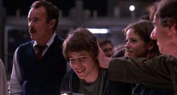 Movie still from “WarGames” (1983), directed by John Badham – A young man smiling while a young woman holds his hand to his ear; Medium shot, Over the shoulder angle