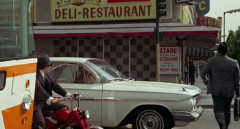 Movie still from “Watermelon Man” (1970), directed by Melvin Van Peebles – A white car parked on the side of the road next to a red motorcycle; Wide shot, Low angle