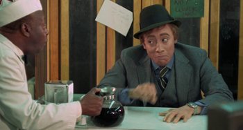 Movie still from “Watermelon Man” (1970), directed by Melvin Van Peebles – A man wearing a suit and a hat sitting at a table; Medium shot, Over the shoulder angle