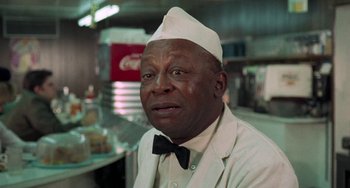Movie still from “Watermelon Man” (1970), directed by Melvin Van Peebles – An older man wearing a white hat and a black bow tie; Close Up shot, Over the shoulder angle