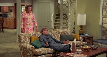 Movie still from “Watermelon Man” (1970), directed by Melvin Van Peebles – An older woman standing next to an older man on a couch; Wide shot, High angle