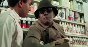 Movie still from “Watermelon Man” (1970), directed by Melvin Van Peebles – A man wearing a hat and sunglasses sitting in a store; Close Up shot, Over the shoulder angle