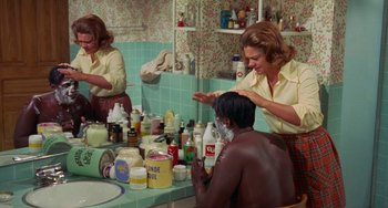 Movie still from “Watermelon Man” (1970), directed by Melvin Van Peebles – Two women are in a bathroom with a man in the background; Medium shot, Over the shoulder angle