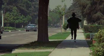 Movie still from “Watermelon Man” (1970), directed by Melvin Van Peebles – A person walking down a sidewalk near a tree; Wide shot, Over the shoulder angle