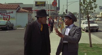 Movie still from “Watermelon Man” (1970), directed by Melvin Van Peebles – Two men talking on the sidewalk of a city street; Medium shot, Over the shoulder angle