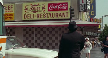 Movie still from “Watermelon Man” (1970), directed by Melvin Van Peebles – A man wearing a black suit and a black hat standing in front of a restaurant; Wide shot, Low angle