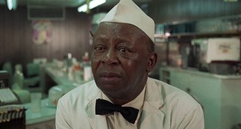 Movie still from “Watermelon Man” (1970), directed by Melvin Van Peebles – An older man wearing a hat and a bow tie; Close Up shot, Over the shoulder angle