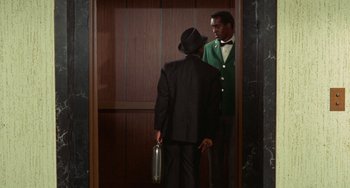 Movie still from “Watermelon Man” (1970), directed by Melvin Van Peebles – A man in a suit standing next to another man in an elevator; Medium shot, Low angle