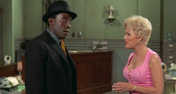 Movie still from “Watermelon Man” (1970), directed by Melvin Van Peebles – A man in a suit and a woman in a pink dress talking; Medium shot, Over the shoulder angle