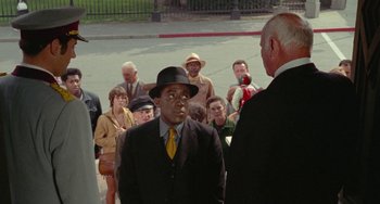 Movie still from “Watermelon Man” (1970), directed by Melvin Van Peebles – A group of people standing around a man wearing a suit and tie; Medium shot, Over the shoulder angle