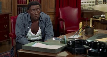 Movie still from “Watermelon Man” (1970), directed by Melvin Van Peebles – A man sitting at a desk in front of a camera; Medium shot, High angle