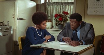 Movie still from “Watermelon Man” (1970), directed by Melvin Van Peebles – Two people sitting at a table in front of each other; Medium shot, Over the shoulder angle
