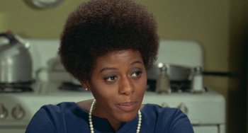 Movie still from “Watermelon Man” (1970), directed by Melvin Van Peebles – An african american woman wearing a blue shirt and pearl necklace; Close Up shot, Over the shoulder angle