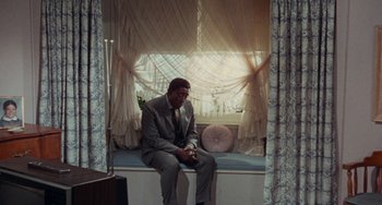 Movie still from “Watermelon Man” (1970), directed by Melvin Van Peebles – A man in a suit sitting on a window sill; Wide shot, High angle