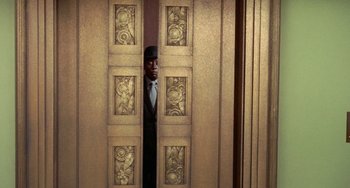 Movie still from “Watermelon Man” (1970), directed by Melvin Van Peebles – A man in a suit and tie standing in front of an open door; Wide shot, Low angle
