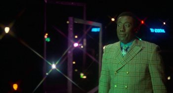 Movie still from “Watermelon Man” (1970), directed by Melvin Van Peebles – A man wearing a suit and tie standing in front of a building; Medium shot, Over the shoulder angle