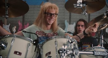 Movie still from “Wayne's World” (1992), directed by Penelope Spheeris – A man with long blonde hair sitting at a drum set; Medium shot, Over the shoulder angle
