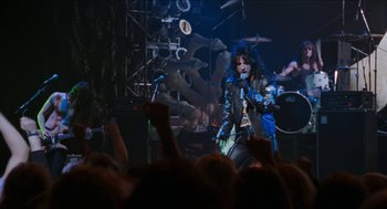 Movie still from “Wayne's World” (1992), directed by Penelope Spheeris – A man in blue jacket holding a microphone on a stage; Wide shot, Over the shoulder angle