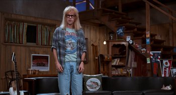Movie still from “Wayne's World” (1992), directed by Penelope Spheeris – A man with long blonde hair and glasses standing on a couch; Wide shot, Low angle