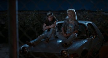 Movie still from “Wayne's World” (1992), directed by Penelope Spheeris – A couple of people sitting on top of a car at night; Wide shot, High angle