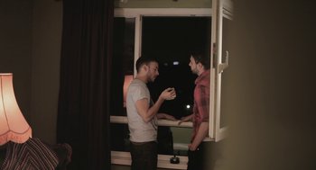 Movie still from “Weekend” (2011), directed by Andrew Haigh – Two young men standing in front of a window at night; Medium shot, Over the shoulder angle