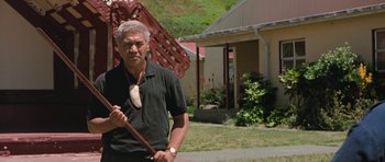 Movie still from “Whale Rider” (2002), directed by Niki Caro – An older man holding a wooden stick in front of a building; Medium shot, Low angle