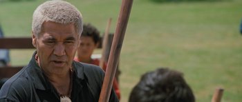 Movie still from “Whale Rider” (2002), directed by Niki Caro – An older man holding a wooden stick in front of a group of people; Close Up shot, Over the shoulder angle