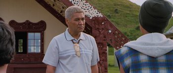 Movie still from “Whale Rider” (2002), directed by Niki Caro – A man wearing a necklace standing in front of a building; Medium shot, Over the shoulder angle