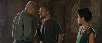 Movie still from “Whale Rider” (2002), directed by Niki Caro – A man standing next to another man in front of a wooden wall; Medium shot, Over the shoulder angle