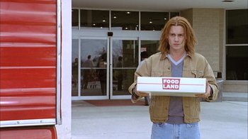 Movie still from “What's Eating Gilbert Grape” (1993), directed by Lasse Hallström – A man holding a box of food in front of a building; Medium shot, Over the shoulder angle
