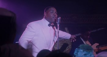 Movie still from “What's Love Got to Do with It” (1993), directed by Brian Gibson – A man in a white suit is singing into a microphone; Medium shot, Low angle