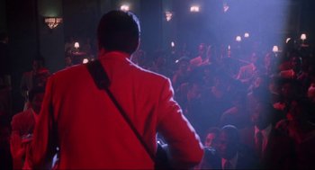 Movie still from “What's Love Got to Do with It” (1993), directed by Brian Gibson – A man in a red jacket is holding a guitar; Wide shot, Over the shoulder angle