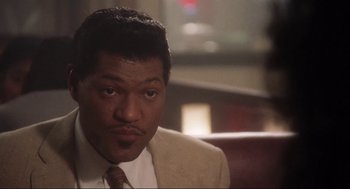 Movie still from “What's Love Got to Do with It” (1993), directed by Brian Gibson – A man sitting in a chair in a room; Close Up shot, Over the shoulder angle